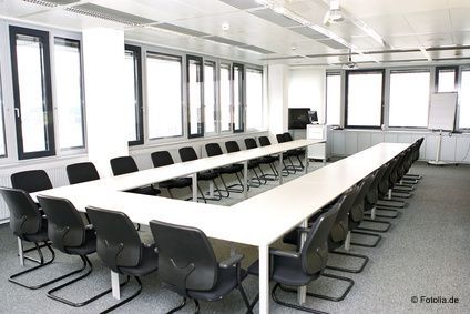 Ein leerer Seminarraum mit hellen Tischen zu einer U-Form aufgestellt und schwarzen Stühlen.