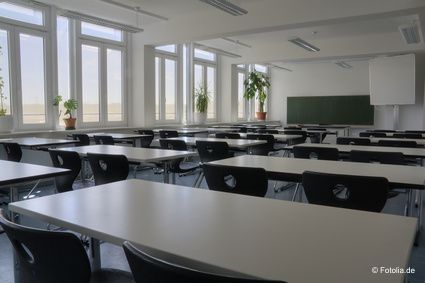 Ein leerer Schulungsraum mit hellen Tischen und schwarzen Stühlen. Aufstellung in drei Reihen mit jeweils mehreren Tischen hintereinander und jeweils zwei Sitzplätzen.
